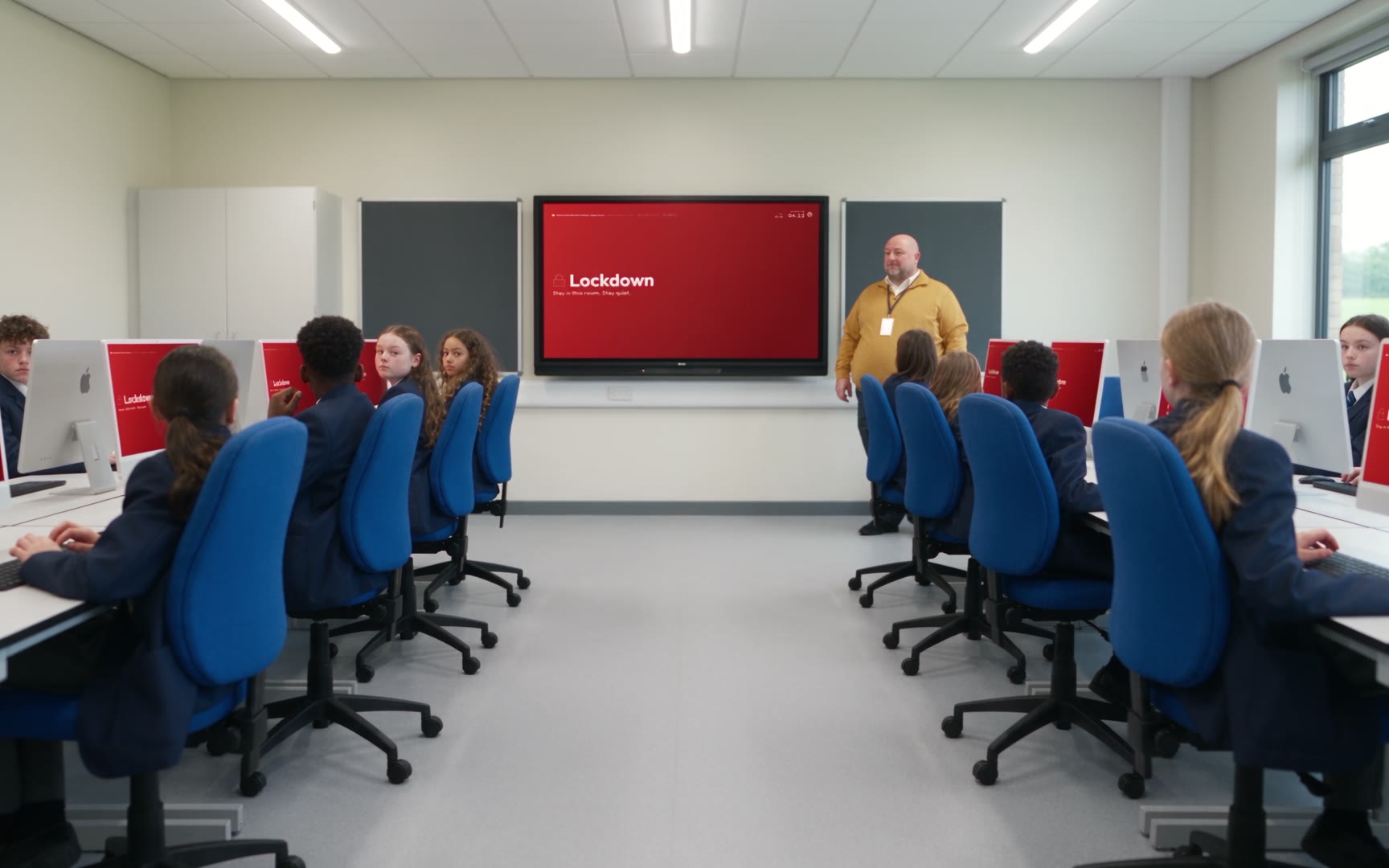 An ICT room in lockdown: the wall display and every iMac screen have turned red with the word Lockdown, pupils at their desks and a member of staff standing by the board