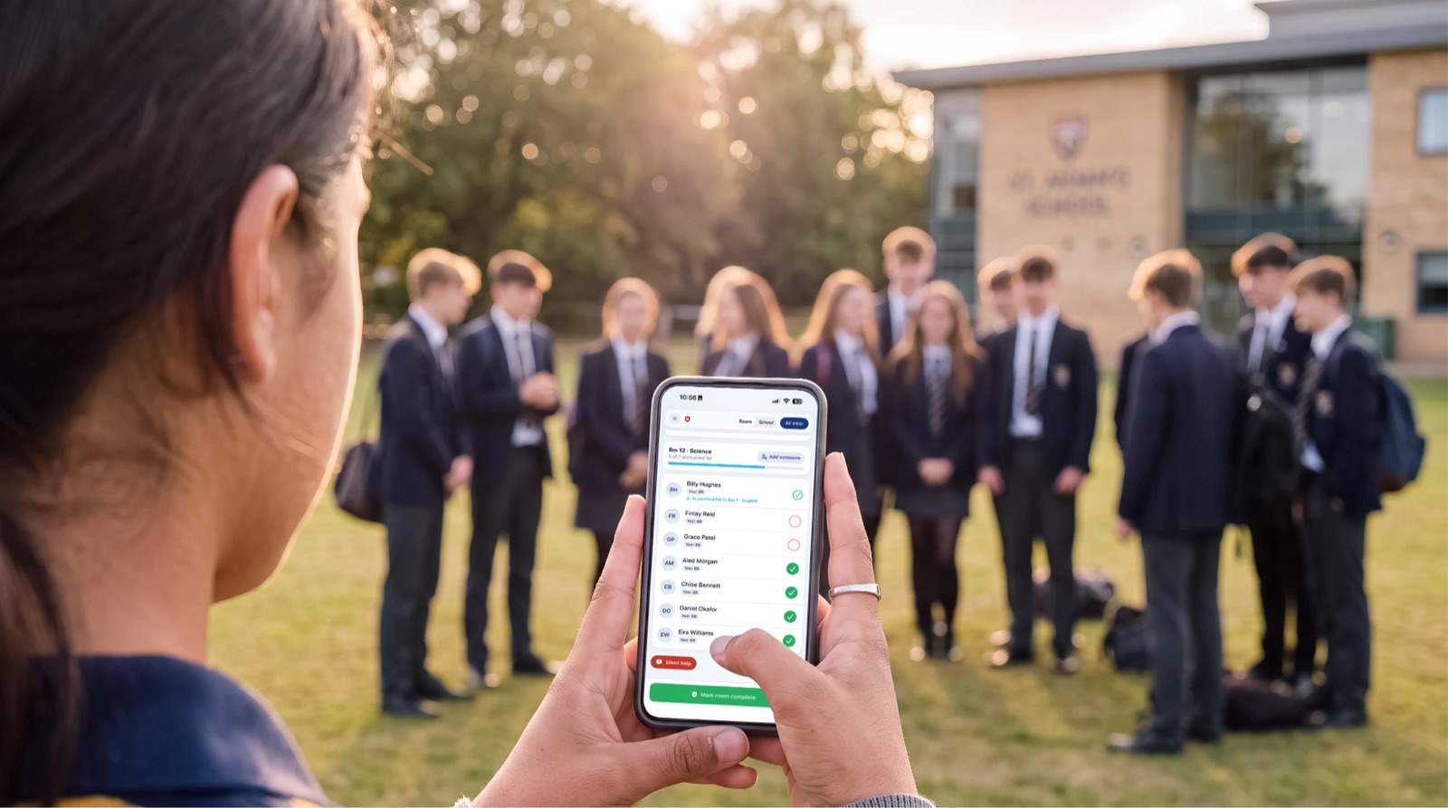 A teacher at the assembly point checking the roll call on her phone, with her class waiting on the field behind her
