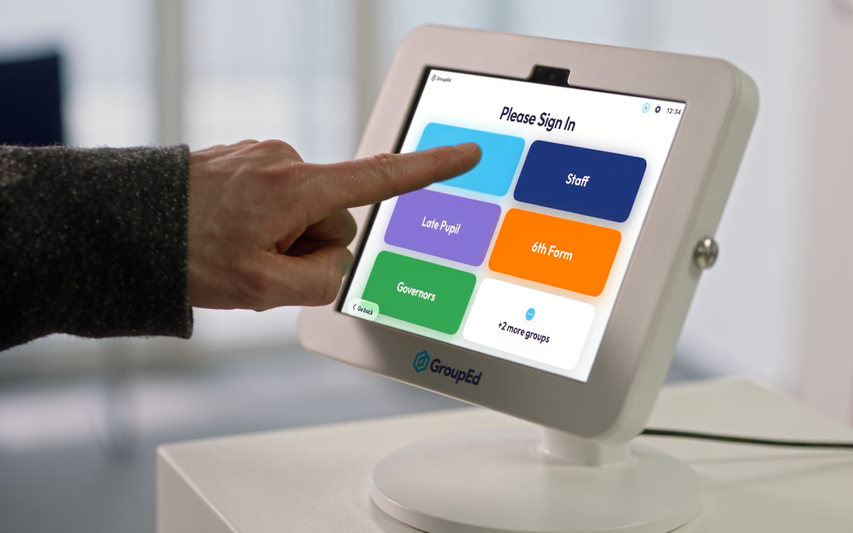 A hand tapping Visitor on the GroupEd sign-in kiosk, running on an iPad at a school reception desk