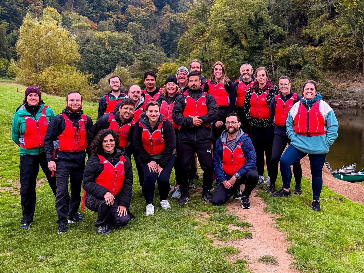 The GroupEd team together on the riverbank in red life jackets, canoes pulled up behind them